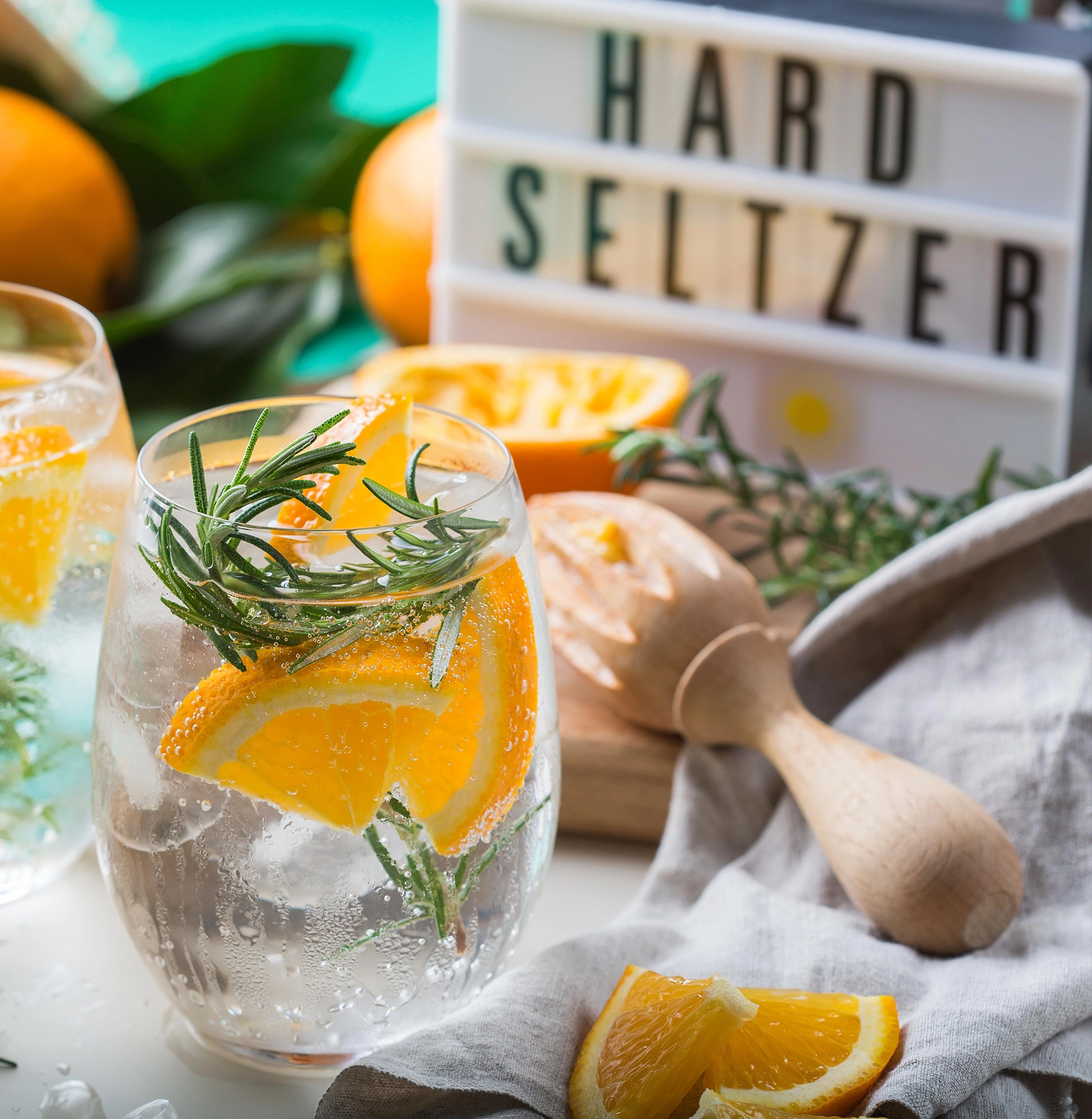 Glas mit Hard Seltzer, Orangenscheiben und Rosmarin auf einem Tisch mit frischen Zitrusfrüchten und dekorativem Hard-Seltzer-Schriftzug.